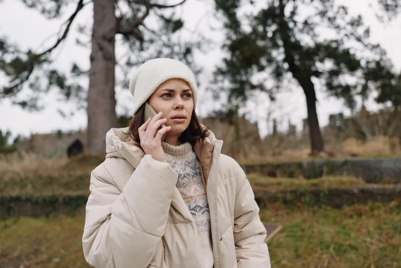 Woman in a light winter coat talks on a mobile phone outdoors in a park, surrounded by trees and open space, capturing a candid moment of remote communication in chilly weather.