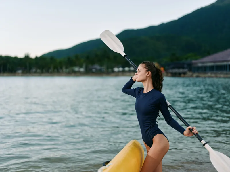 Woman in swimsuit holding paddle stands by yellow kayak on lake with mountains and forest background in summer.