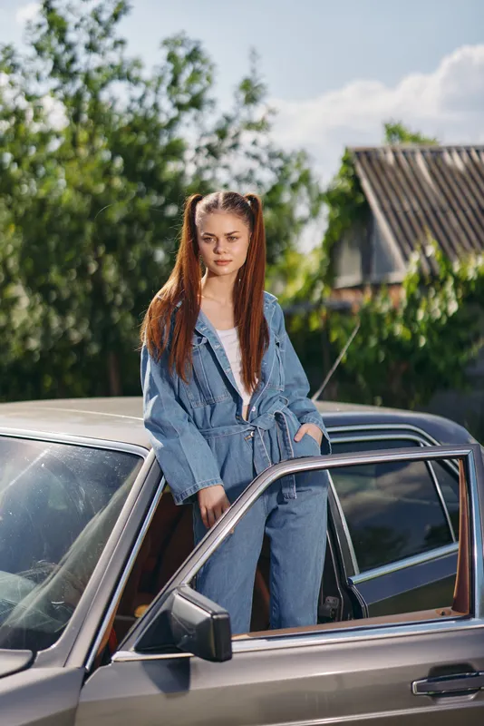 Stylish young woman in denim overalls confidently posing next to a classic car in a sunny outdoor setting, showcasing casual fashion and seasonal vibes.