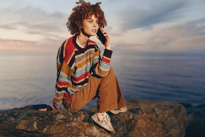 Woman lifestyle outdoors sea sunset, curly hair sweater casual outfit, sitting on rocky coast, thoughtful smile, warm golden light, soft breeze, natural colors, cinematic mood at dusk ocean