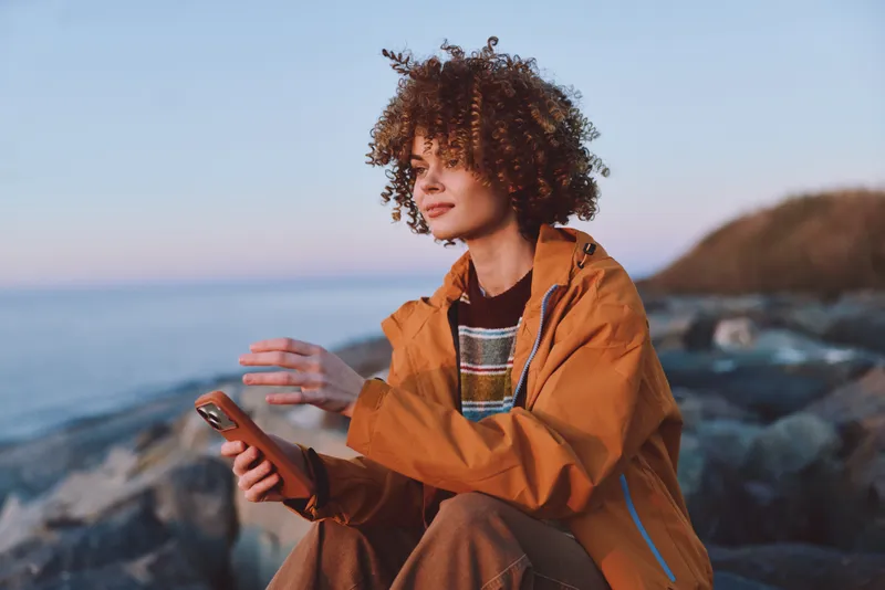 Woman lifestyle seaside smartphone curly jacket ocean rocks, young woman in orange outerwear holds a smartphone on rocky shore, gazing at the horizon, relaxed smile, sunset breeze
