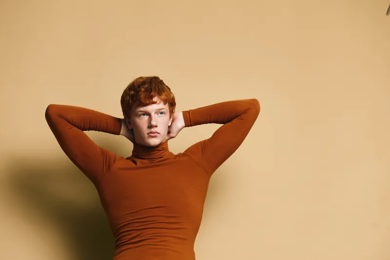 Young man with red hair wearing brown turtleneck posing with hands behind head against beige wall looking away thoughtful and stylish casual look for fashion portrait.