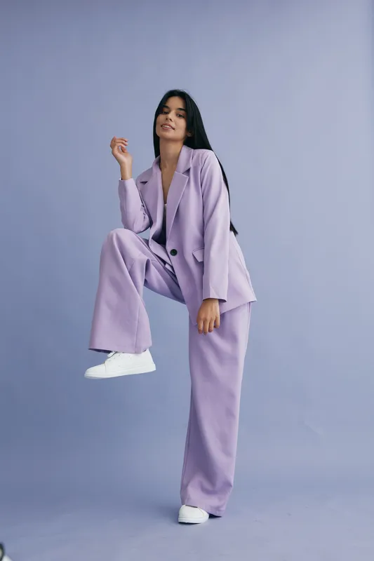 Woman in lilac pantsuit posing confidently against a soft purple background, wearing white sneakers with straight black hair, showcasing modern fashion style.