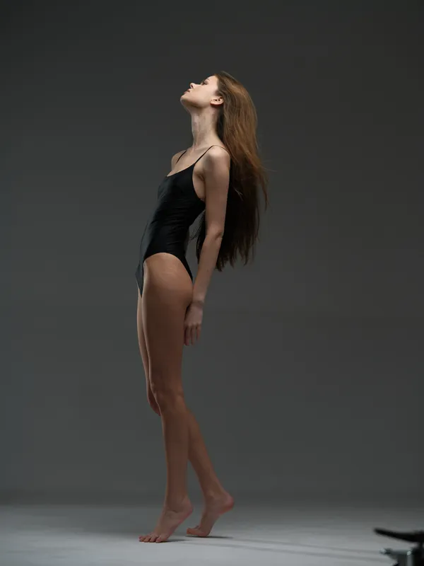 slender woman in black bodysuit standing on tiptoes with head tilted back, minimalist dark background, elegant pose