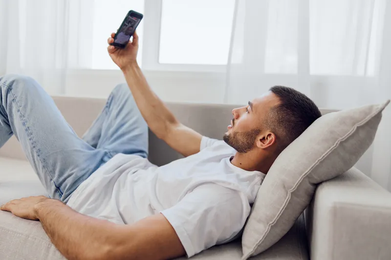 Thoughtful man relaxing on a couch, engaging with his smartphone, reflecting the challenges of technology, work-life blur, and mental health awareness amid daily life.
