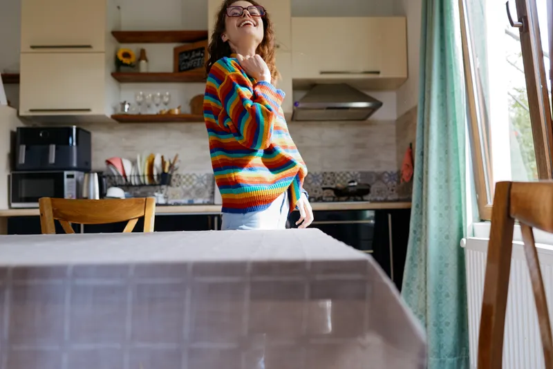 Young woman wearing a colorful striped sweater stands in a bright modern kitchen, laughing with her head tilted back, capturing a warm casual home moment
