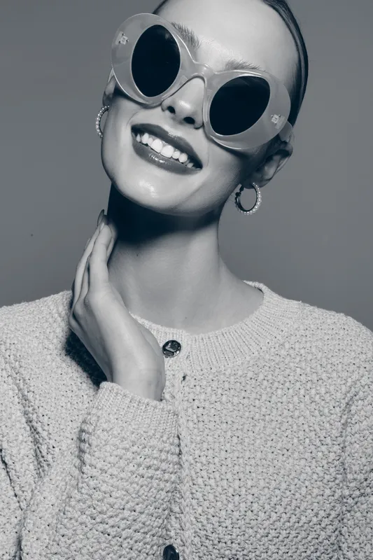 Stylish smiling white woman with pale skin wears large round sunglasses and textured sweater. Glamorous fashion portrait with confident mood in studio black and white photo.