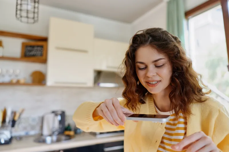 Young woman in a yellow cardigan smiles while serving a dish in a bright home kitchen, capturing a candid moment of daily cooking and warm atmosphere at home