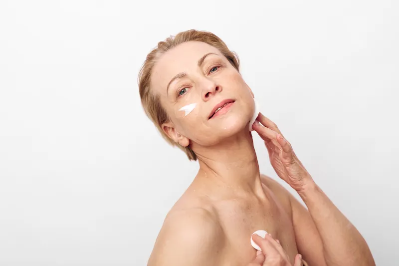 Mature woman with short hair applies face cream on cheeks and neck, showing skincare routine and self-care concept with soft lighting on neutral background for healthy skin.