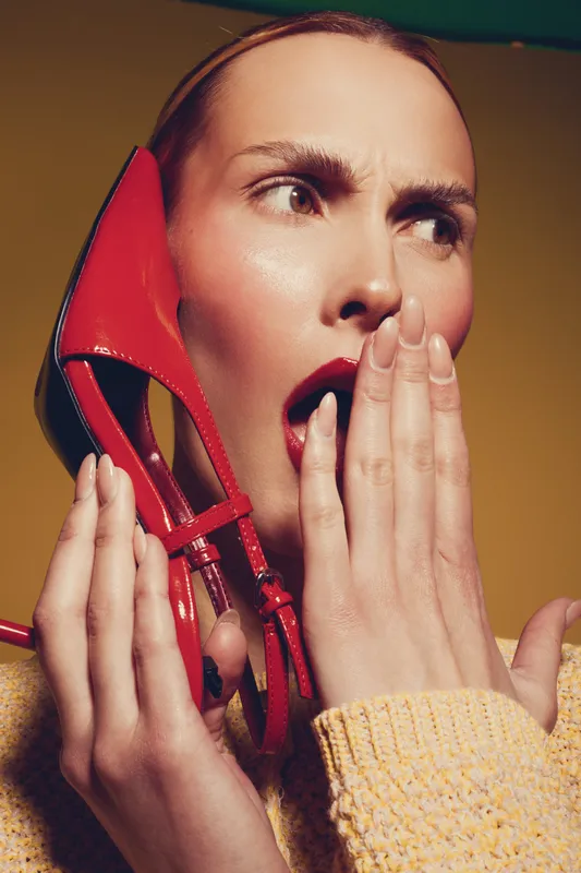 Surprised woman with red lipstick holds red high heel shoe near face, wearing yellow sweater. Studio portrait on yellow background with dramatic expression and fashion concept.