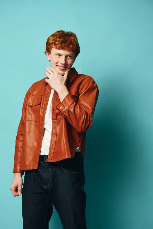 Young man with red hair wearing brown leather jacket and white shirt posing with hand near chin against light blue background smiling confidently.