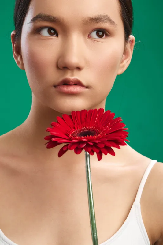 asian woman stands against a green backdrop, serene portrait with a red flower near her chin, soft lighting and smooth skin create a calm beauty aesthetic.