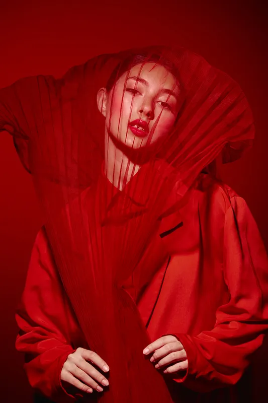 Fashion woman in intense red outfit and dramatic mesh accessory poses with eyes closed on red background. Creative concept of beauty, style, and artistic expression.
