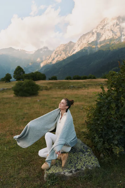 Young woman wrapped in a blanket sitting on a rock in a mountain meadow. Relaxed lifestyle concept with scenic nature background and soft natural light.