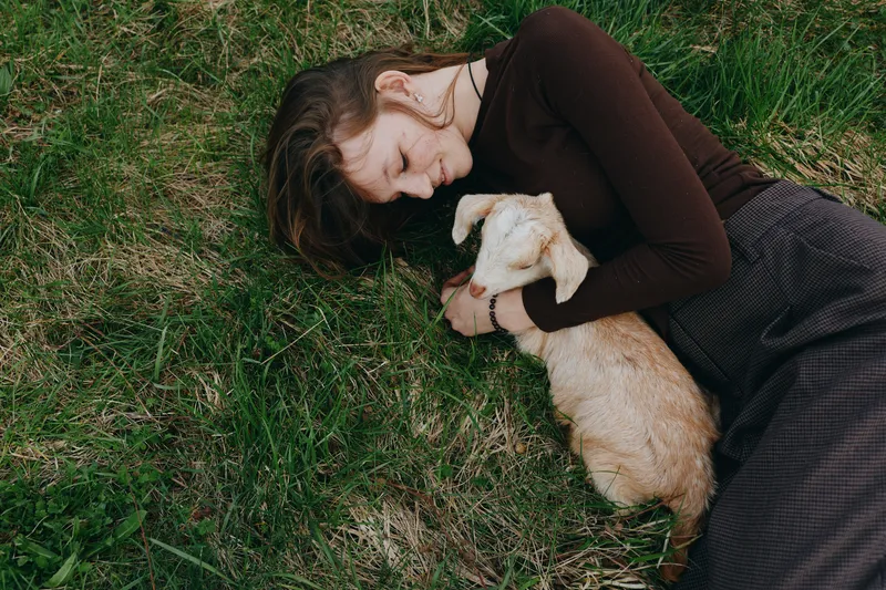 A woman lies on green grass embracing a young goat kid, sharing a tender outdoor moment. Rustic lifestyle, companionship with animals, and quiet affection in nature. A candid, peaceful scene evokes