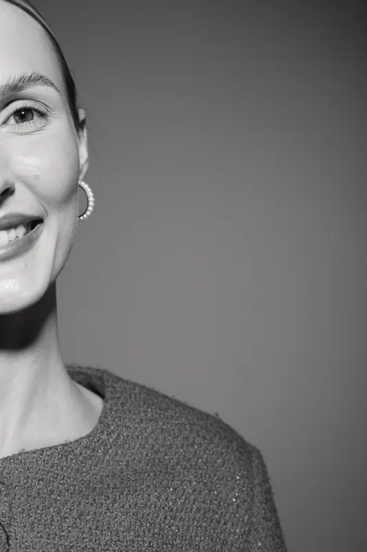 Close-up of a smiling Caucasian woman with fair skin wearing a textured sweater and hoop earrings on grey background.