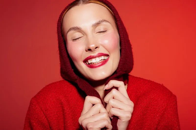 Smiling woman in red knitted hoodie with closed eyes enjoys cozy warmth, isolated on red background. Concept of happiness, comfort, and winter fashion style.
