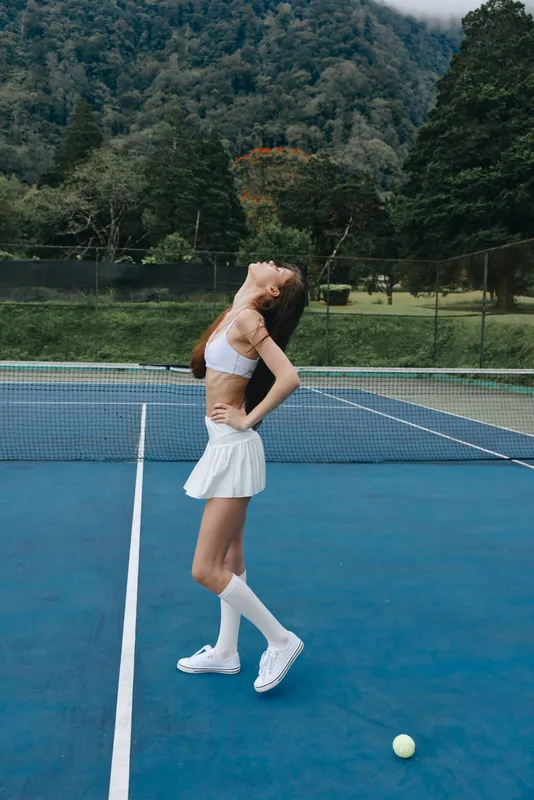 Athletic young woman in a white tennis outfit poses confidently on a court surrounded by green mountains, showcasing emotions of determination and joy