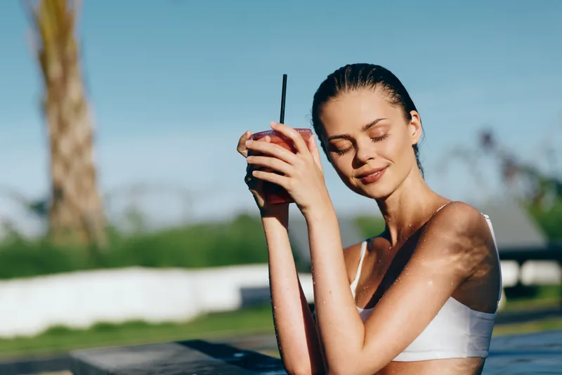 Sunlit woman in a white bikini top sits by a pool, sipping a cold berry smoothie with a straw. Relaxed lifestyle, wellness, and self care in bright, warm daylight.