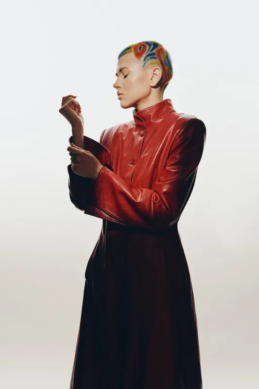 Stylish woman with colorful patterned hair wears a sleek red leather coat, posing thoughtfully against a light neutral background, showcasing unique fashion and bold design.
