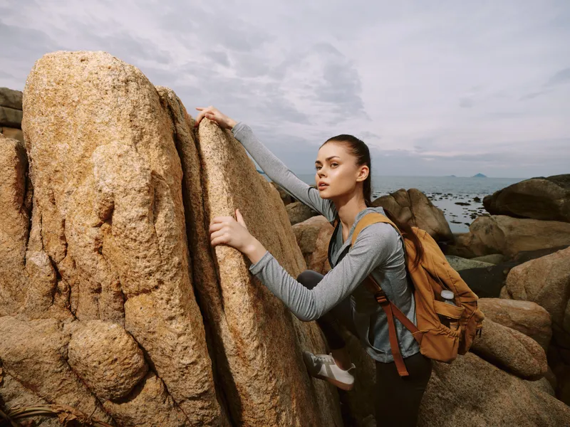 Young woman climbing rocky terrain with a backpack, wearing casual outdoor clothing, expressing determination in a natural landscape