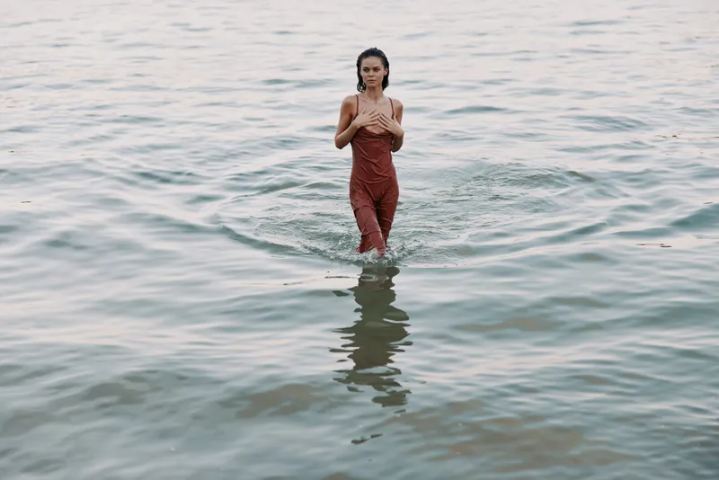 A solitary figure stands in shallow sea water, wearing rust red overalls, hands crossed at chest, while soft light and gentle ripples frame a calm, contemplative mood.