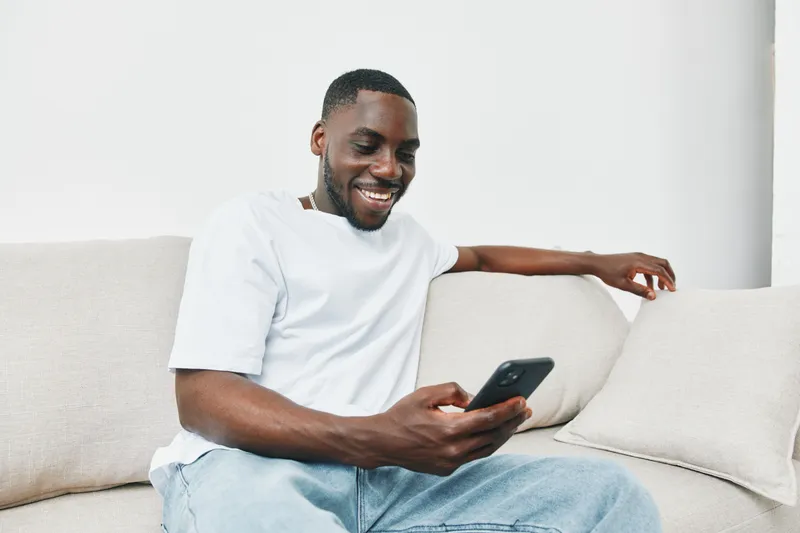 Smiling Black man casually browsing smartphone at home on a cozy couch in a relaxed atmosphere, enjoying leisure time and technology