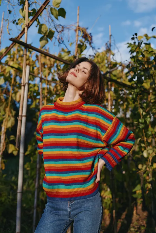 Woman in her mid twenties wearing a colorful striped sweater and jeans outdoors in a sunlit vineyard, smiling and leaning back in a casual outfit for lifestyle fashion.