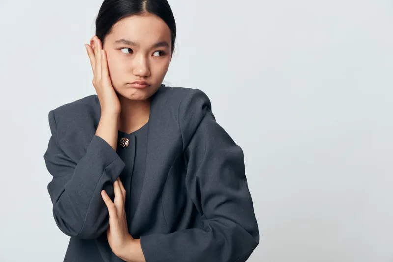 Young woman in dark gray jacket covering ear and looking away with worried expression on light background. Portrait of frustrated female with hand near head in studio for anxiety or discomfort.