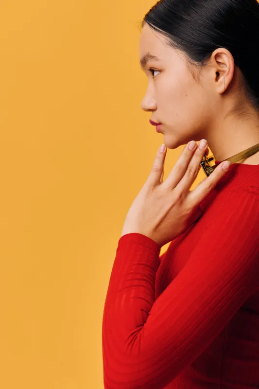 Young woman in red top poses against yellow background with thoughtful expression, hand near neck, side profile, minimal makeup and sleek black hair tied back, fashion portrait.