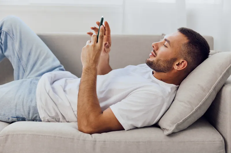 Relaxed man using smartphone while lounging on the sofa, displaying a modern lifestyle and comfort in a cozy indoor setting. Perfect for showcasing leisure and technology.