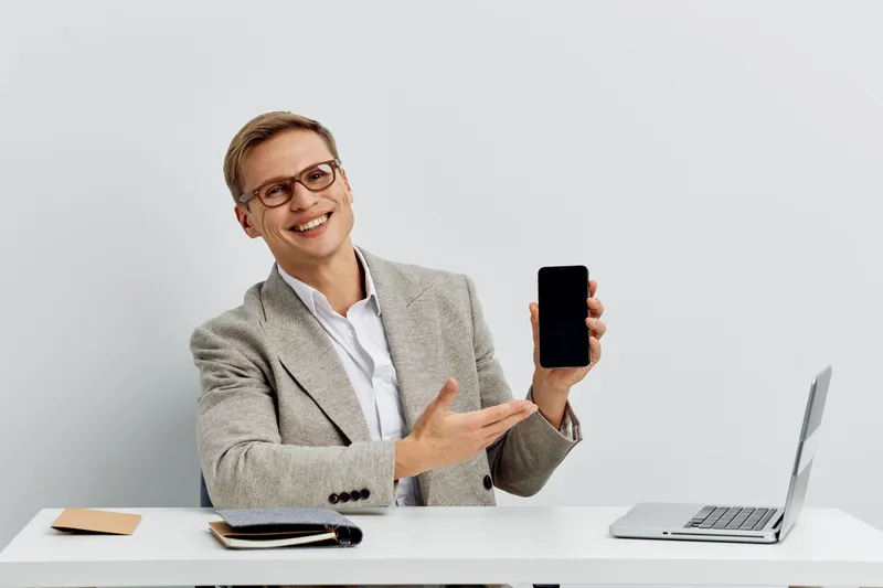 Smiling woman in office attire holding smartphone and showing it to the camera, sitting at a desk with laptop and notebooks, white background. Technology, communication, modern, professional, business