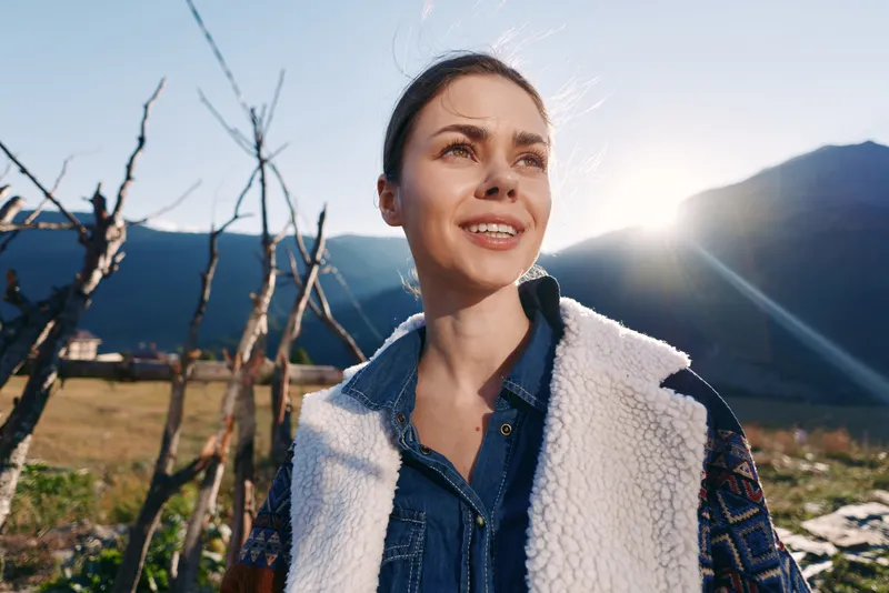 Woman portrait in mountains at sunrise smile outdoors with sunlight on face, casual denim shirt and shearling vest, scenic nature background and peaceful outdoor travel mood