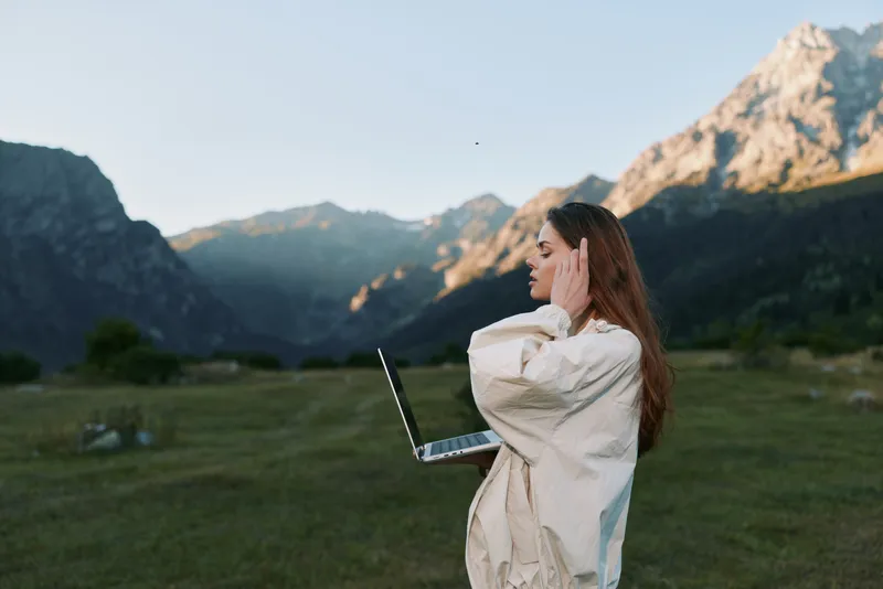 A young woman stands in a mountain meadow, wearing a light jacket and using a laptop. She appears focused and calm, enjoying fresh air while staying connected to work from a serene outdoor setting.