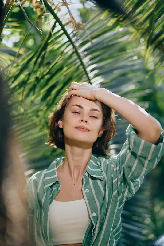 Portrait of a calm woman basking in sunlight beneath palm leaves, wearing a green striped shirt and white top.