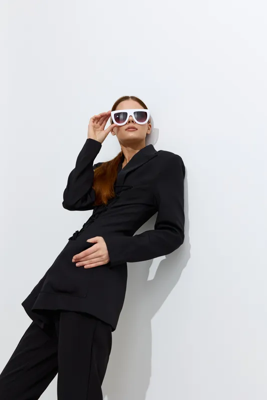 Fashion woman dressed in sleek black outfit poses confidently with white sunglasses against minimalist white background, showcasing modern style and sophistication.