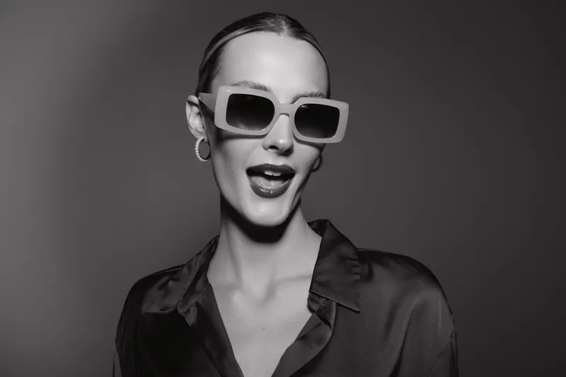 Young white woman with light skin wears oversized sunglasses and silk shirt posing with open mouth in studio. Concept for fashion, style, confidence, and beauty portrait.