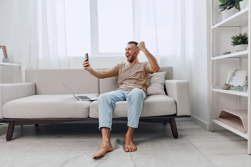 Excited man celebrating success while using a smartphone, casual attire, bright living room ambiance, modern decor, showcasing emotions of happiness and achievement.