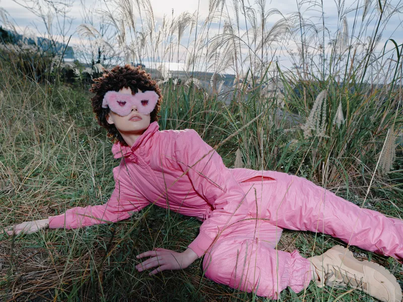 Pink jumpsuit worn by a masked person lying in tall grass outdoors, creating a fashion cosplay vibe with a dramatic pose in nature and vivid color.