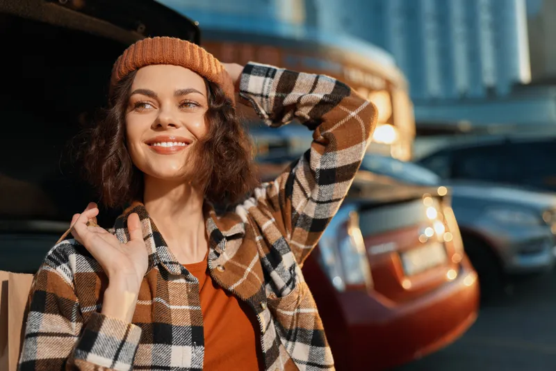 Woman with a warm smile in candid golden light by her car, urban beanie style and cozy plaid shirt. Authenticity, mindful living and emotional storytelling in a lifestyle portrait.
