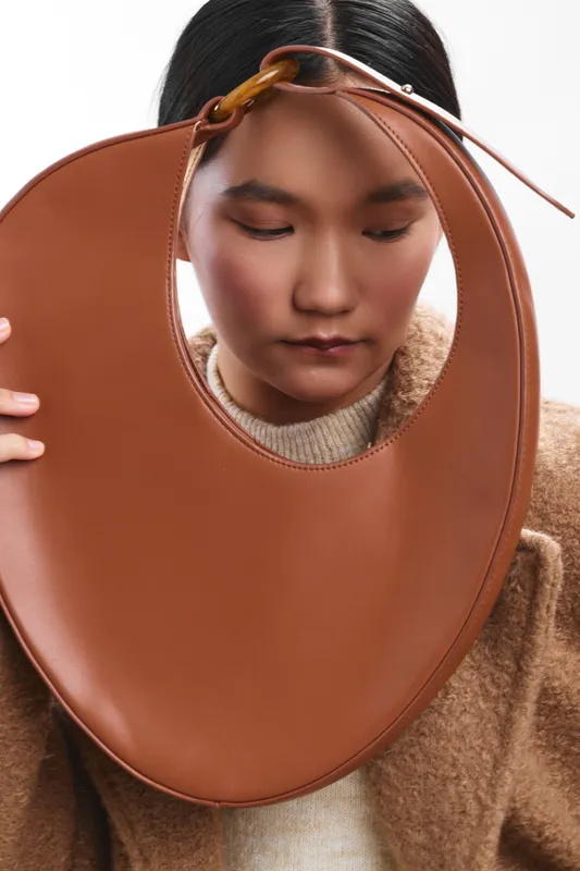 asian woman wearing a cozy sweater gazes downward as a large brown circular bag rests near her face, creating a minimal fashion scene highlighting texture, style, and contemplative mood.