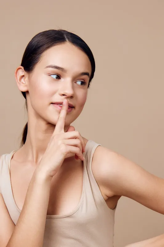 Young woman with a neutral expression, wearing a natural toned tank top, holding a finger to her lips against a soft beige background, promoting serenity and calmness