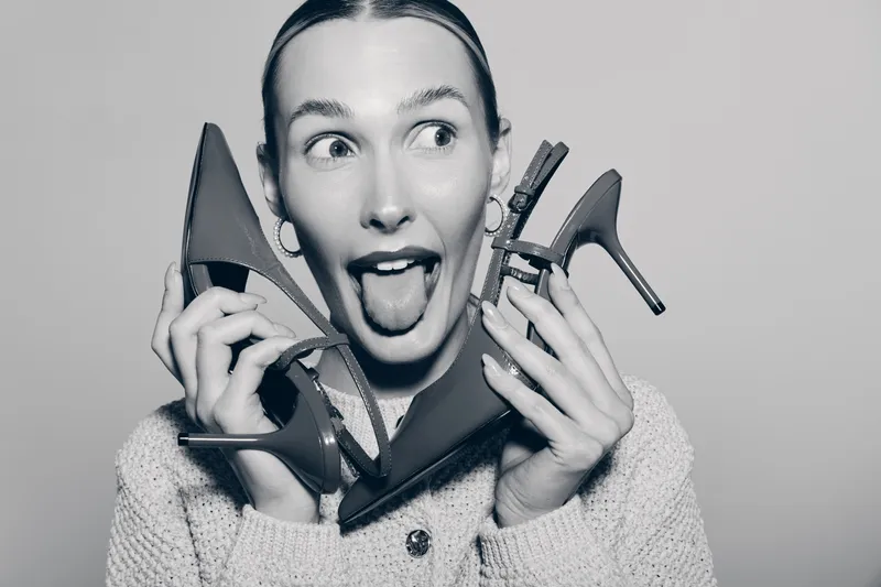 Caucasian woman with fair skin holds two high heel shoes near her face, making playful expression with tongue out, isolated on plain background.