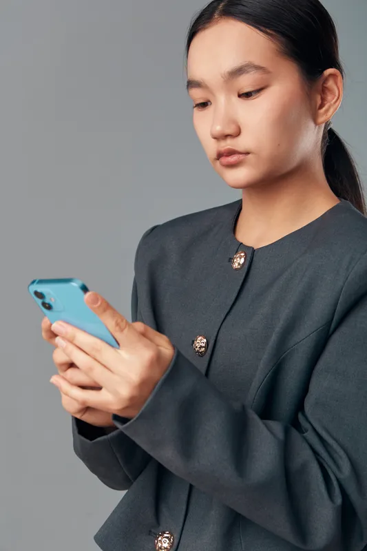 Portrait of young woman using smartphone with focus and concentration against gray background. Asian female dressed in dark outfit holding mobile phone for communication or social media.