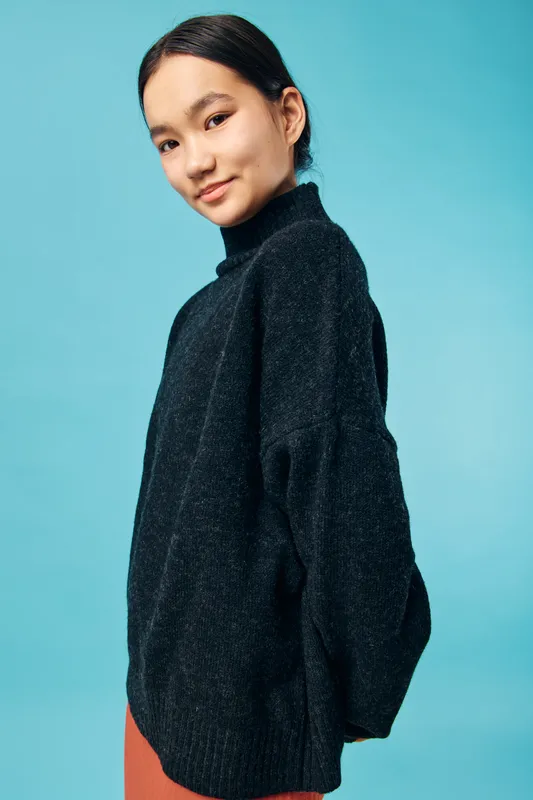 Young woman wearing oversized black sweater posing sideways on blue background, casual fashion style, soft expression and natural makeup, modern portrait in studio lighting
