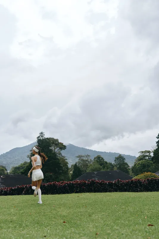 Young woman running on a green lawn, dressed in white athletic wear, displaying joyfulness amidst a serene outdoor backdrop of mountains and cloudy skies