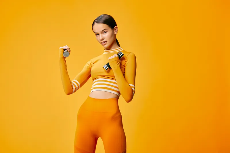 Young fit woman in matching orange activewear holding small dumbbells, posing confidently in a bright orange studio environment. Minimal seamless background, energetic fitness concept.