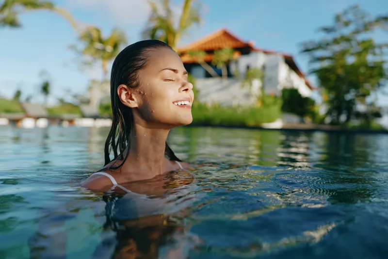 Smiling woman enjoying a sunlit pool, body relaxed as clear water surrounds her, tropical resort setting, warm breeze, leisure and wellness vibe, bright inviting mood.
