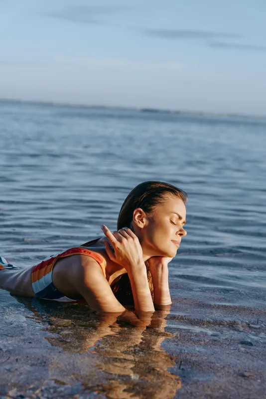 Woman at beach in shallow ocean water, waves softly in background, relaxed kneeling portrait, sunlight highlights wet hair and skin, calm evening, quiet mood, natural colors