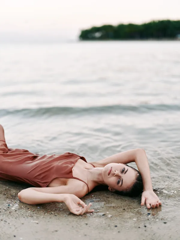 A woman in a pink dress lies on the sandy beach at the water's edge, with soft waves in the background and a calm, contemplative mood.